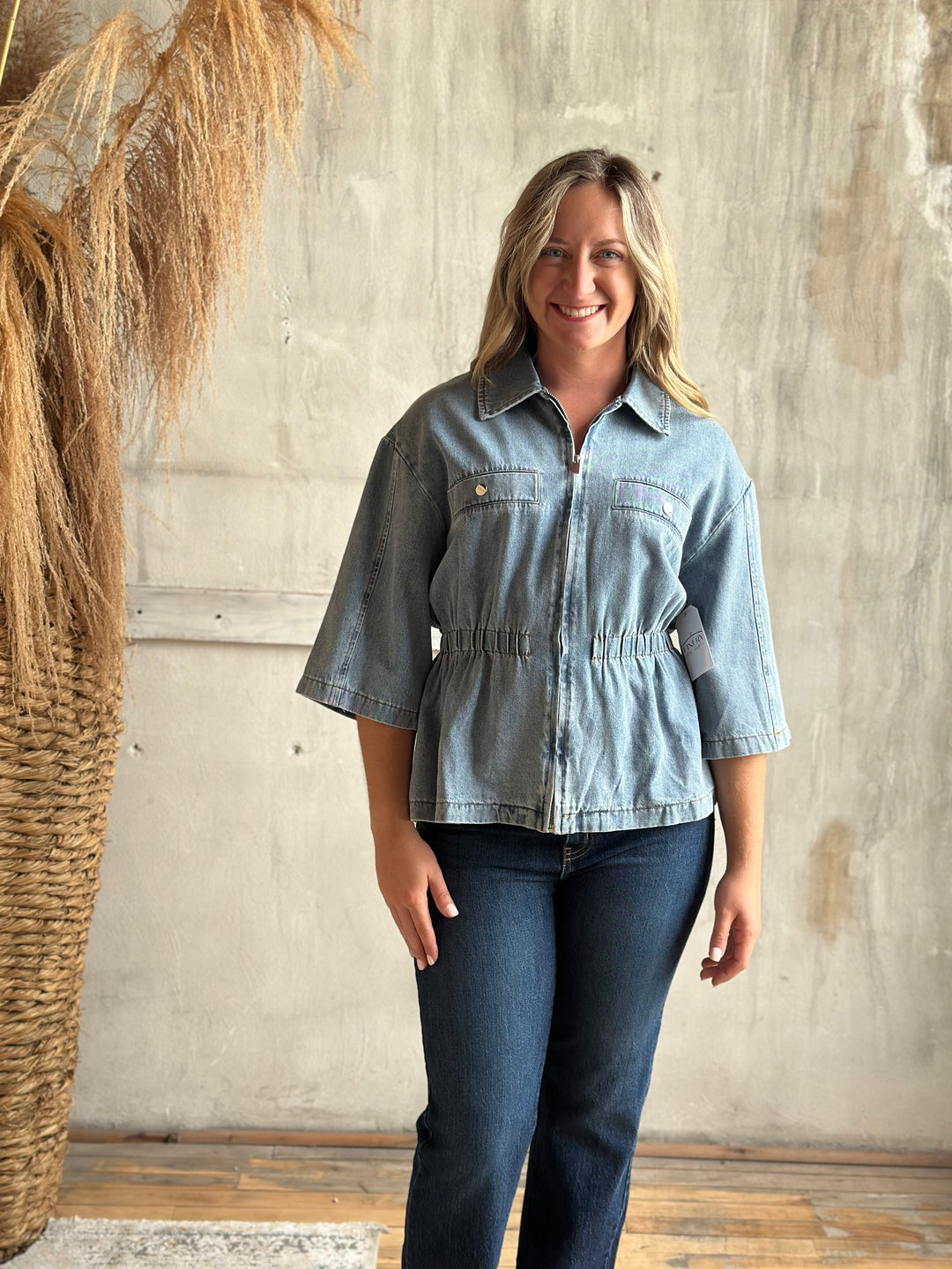 A woman modeling a light-wash blue denim jacket with three-quarter sleeves and a gathered waist, paired with dark blue jeans, standing against a textured concrete wall next to a dried pampas grass arrangement.