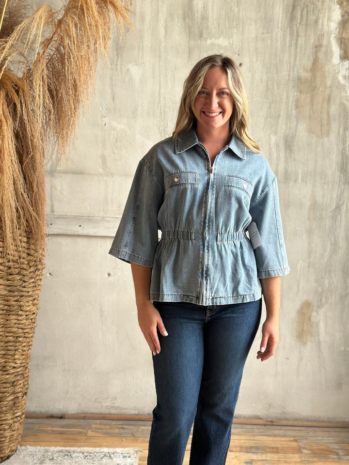 A woman modeling a light-wash blue denim jacket with three-quarter sleeves and a gathered waist, paired with dark blue jeans, standing against a textured concrete wall next to a dried pampas grass arrangement.