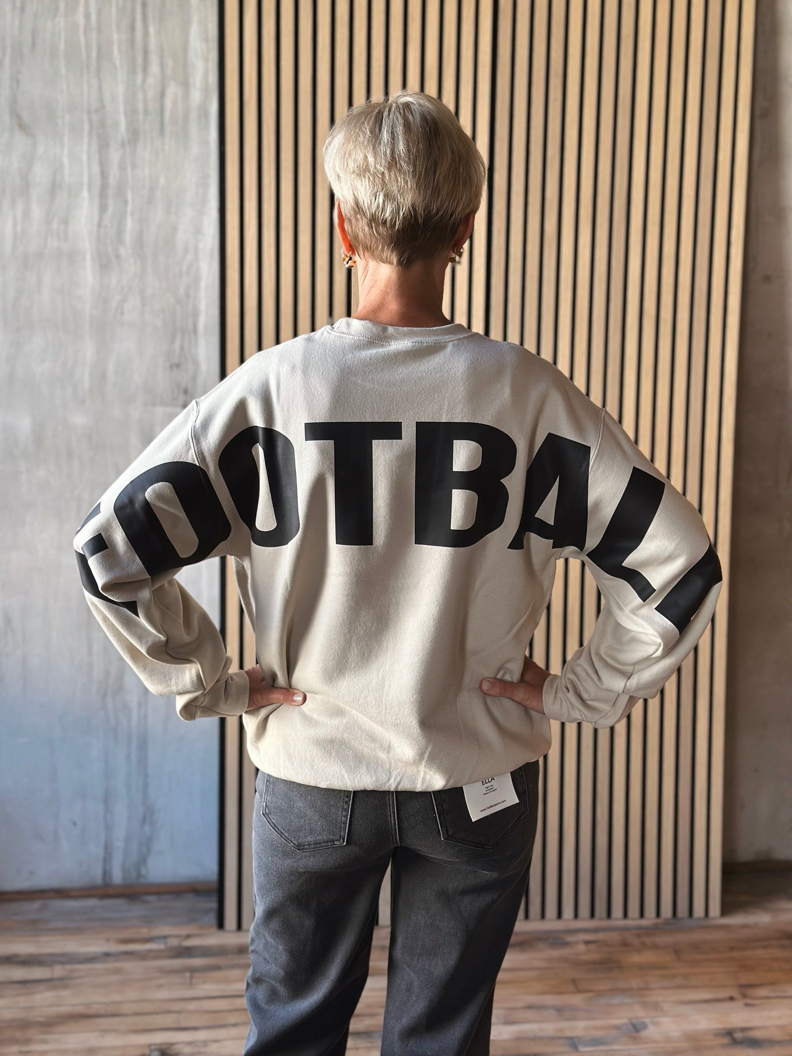 Person wearing a white sweatshirt with 'FOOTBALL' printed on the back, standing against a wooden paneled wall.