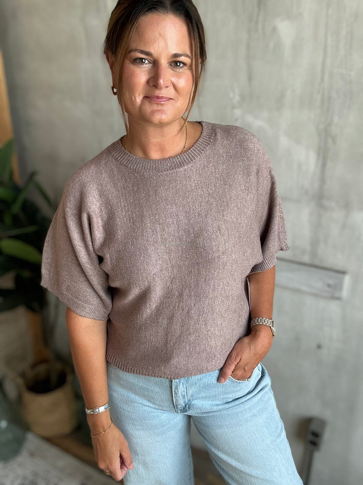 A woman wearing a short-sleeve knit sweater in a dusty taupe-brown color, paired with light blue jeans.