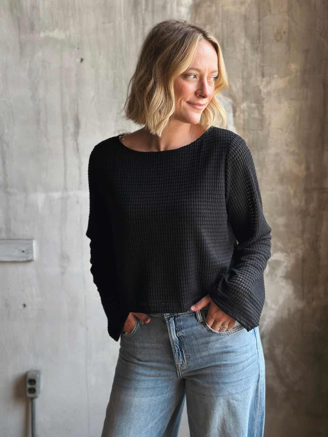 Woman wearing a black sweater and light blue jeans against a concrete wall.