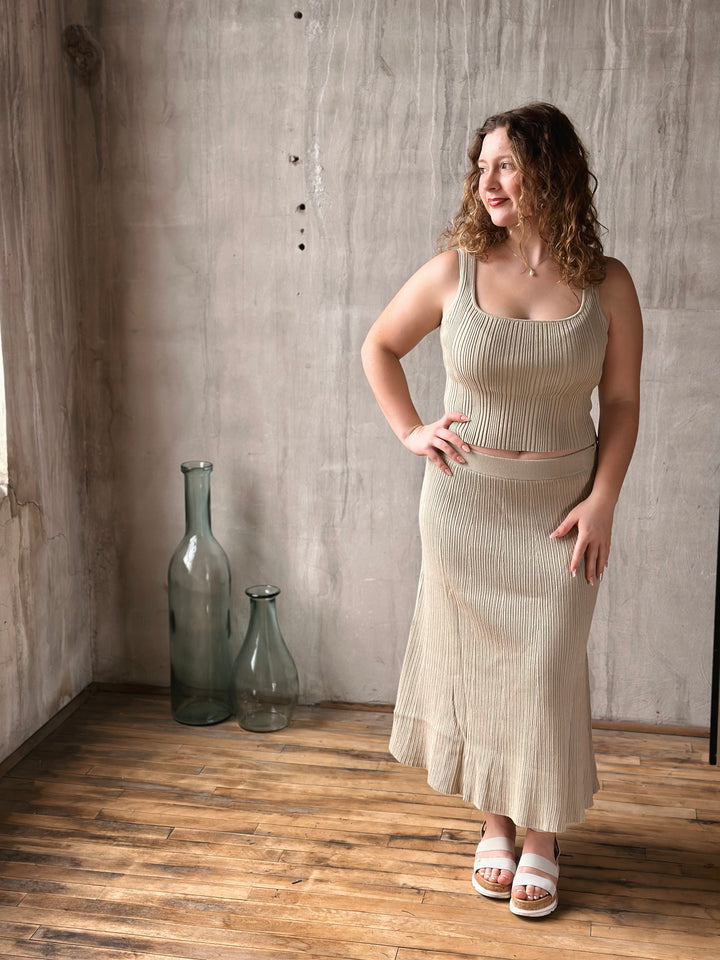 Woman wearing a beige ribbed knit two-piece set standing in a room with wooden floor and textured wall.
