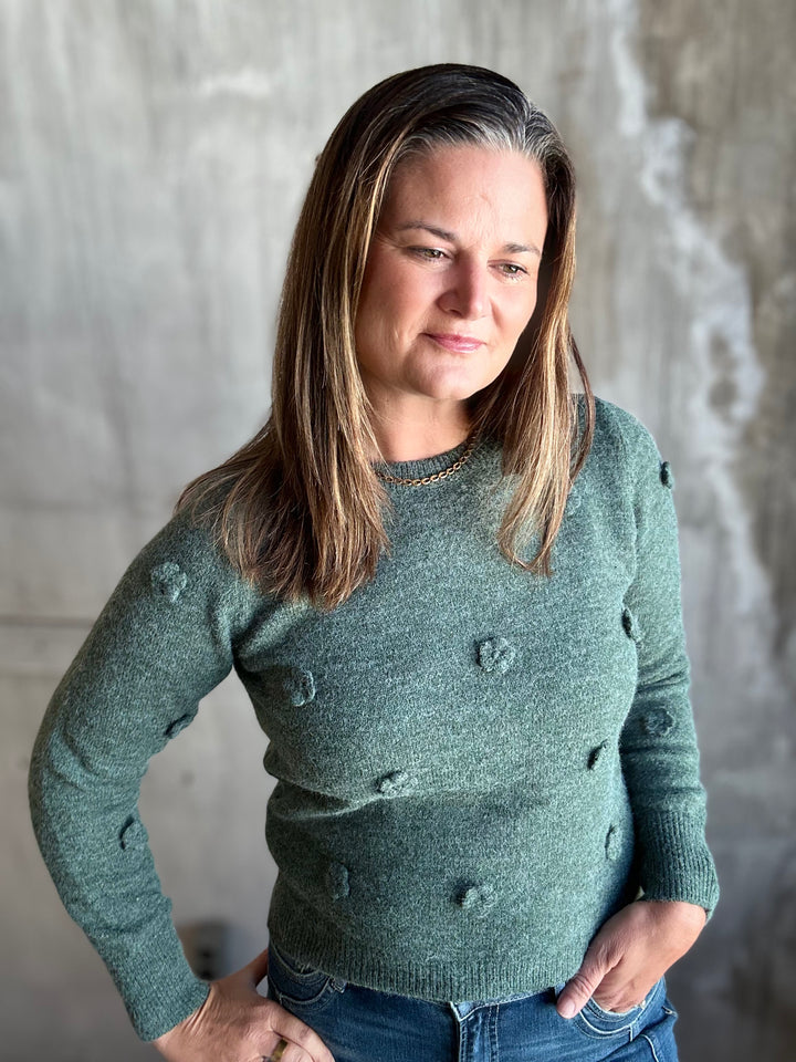 A woman wearing a sage green knit sweater with textured pom-pom details, paired with blue jeans, standing against a textured gray concrete wall.