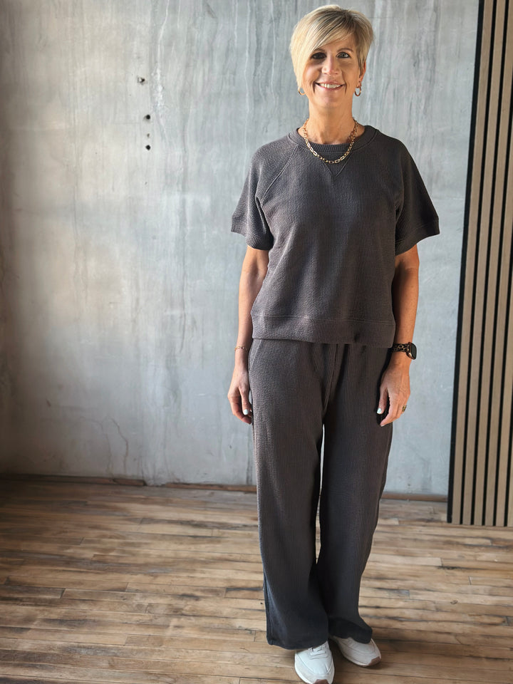 Woman wearing a dark gray outfit standing in a room with wooden flooring and a concrete wall.