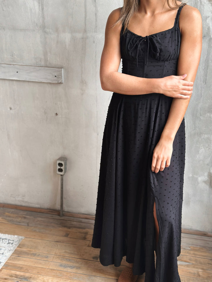 Woman wearing a black dress with a slit in a room with concrete walls and wooden floor.