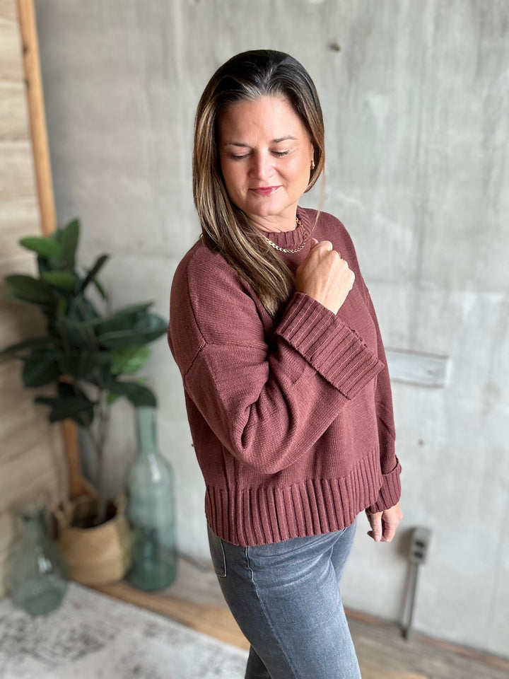 A woman wearing a dusty rose ribbed knit sweater and blue jeans, standing in a room with a concrete wall, plants, and glass bottles.