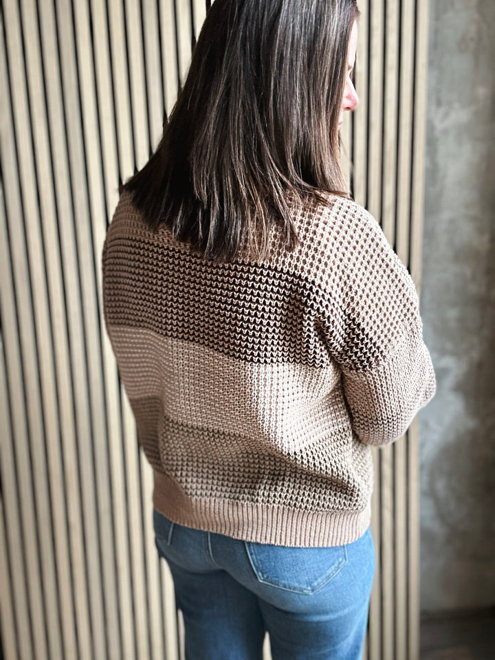 Person wearing a beige textured sweater and blue jeans against a neutral wall.