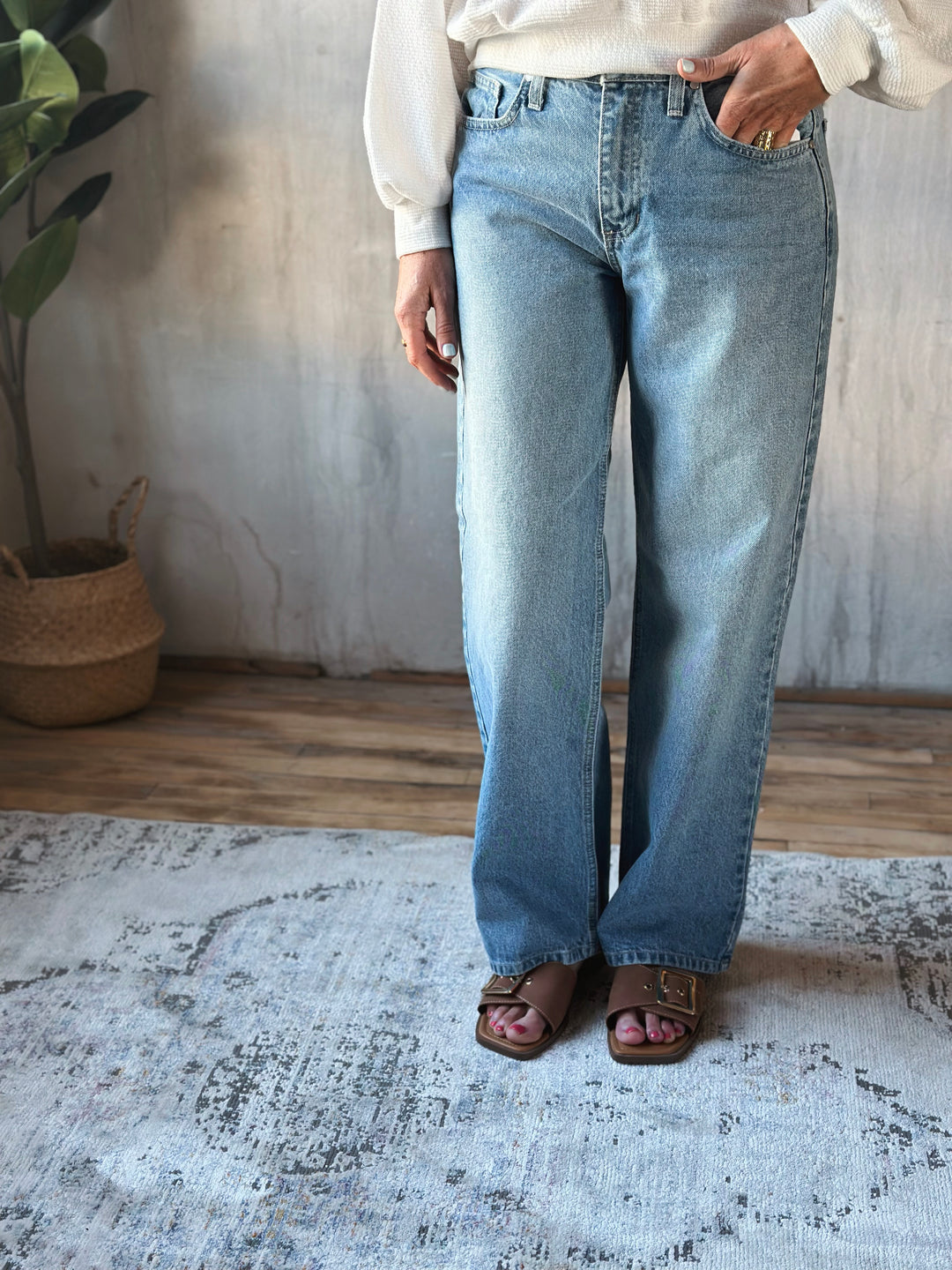 Person wearing light blue jeans standing in a room with a plant and wooden floor.