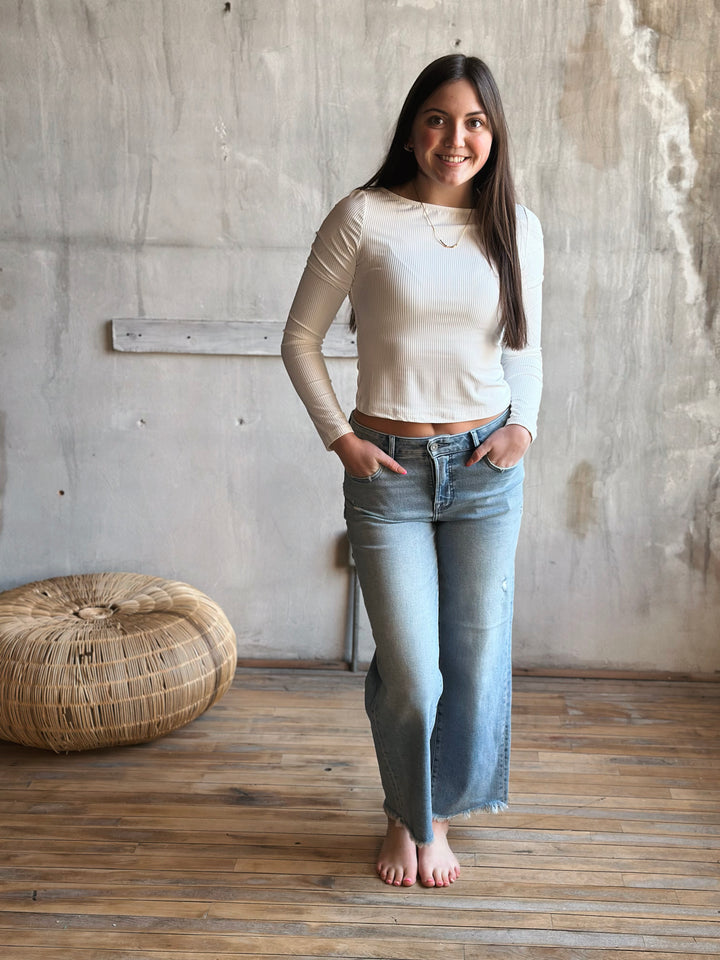 Woman wearing a beige long-sleeve top and light blue jeans standing in a room with wooden flooring and a textured wall.