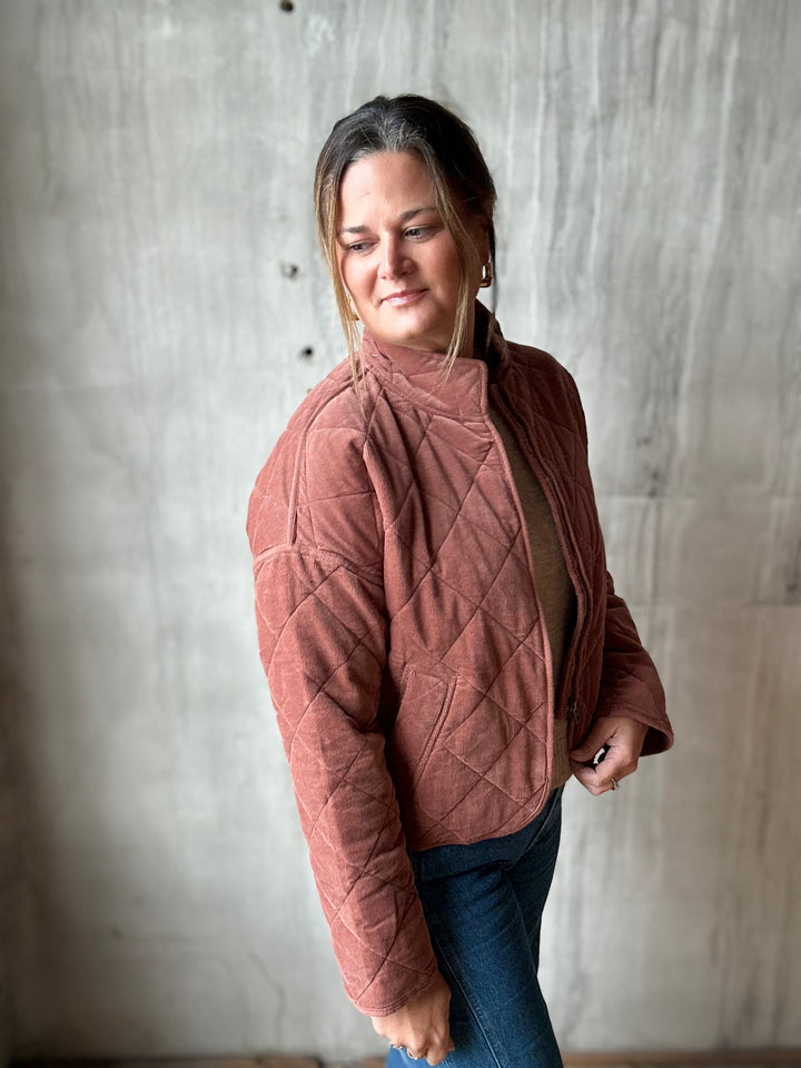 A woman wearing a dusty rose quilted jacket and blue jeans, standing against a textured concrete wall.