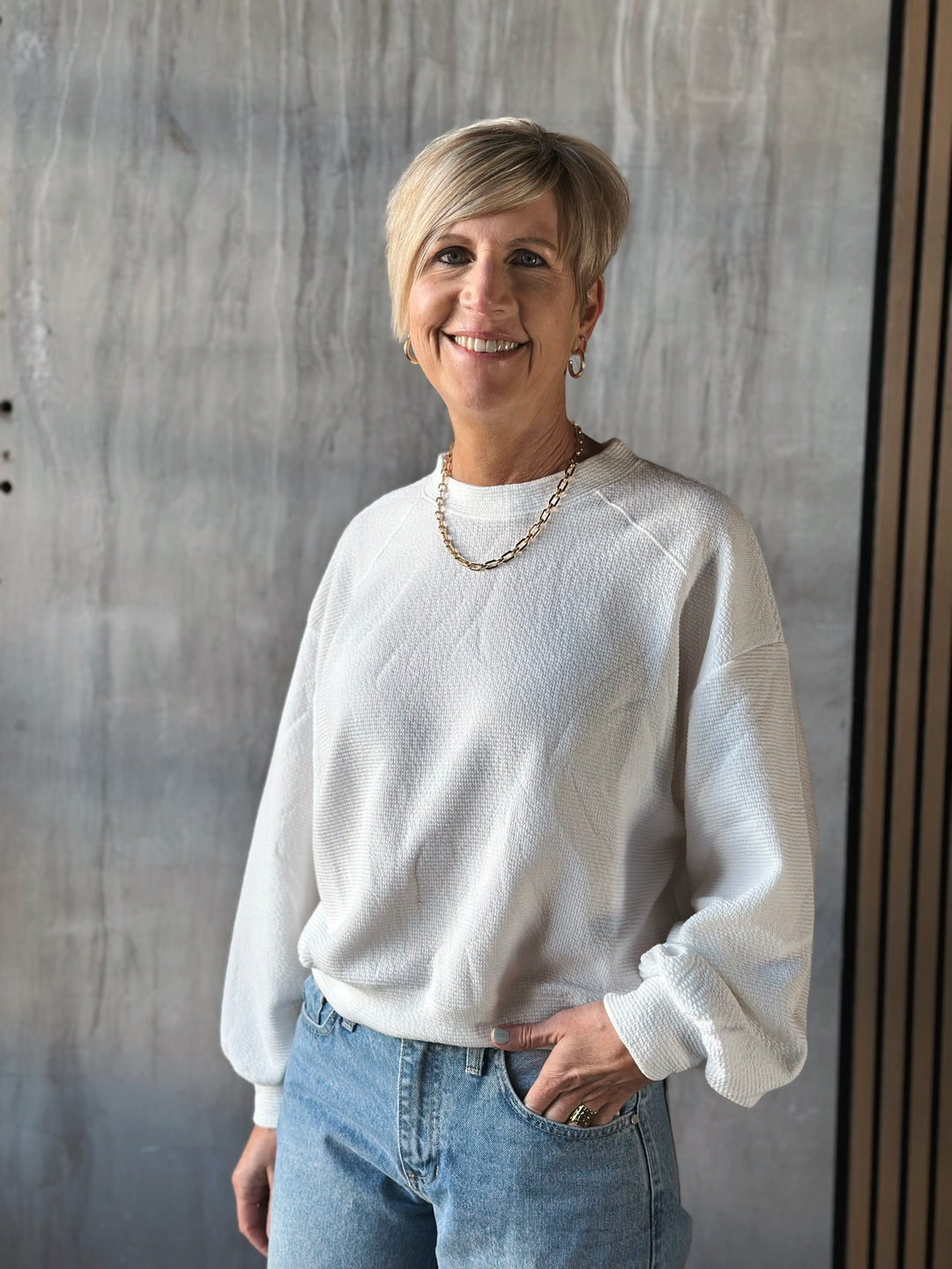 Woman wearing a white sweater and blue jeans standing against a gray wall.