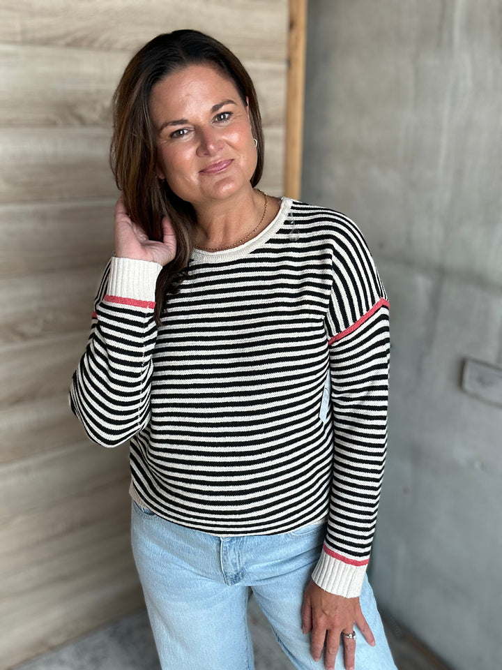 A woman wearing a black and white horizontally striped long-sleeve sweater with pink accent stripes on the cuffs and upper sleeve, paired with light blue jeans, standing in front of a wood-paneled wall.