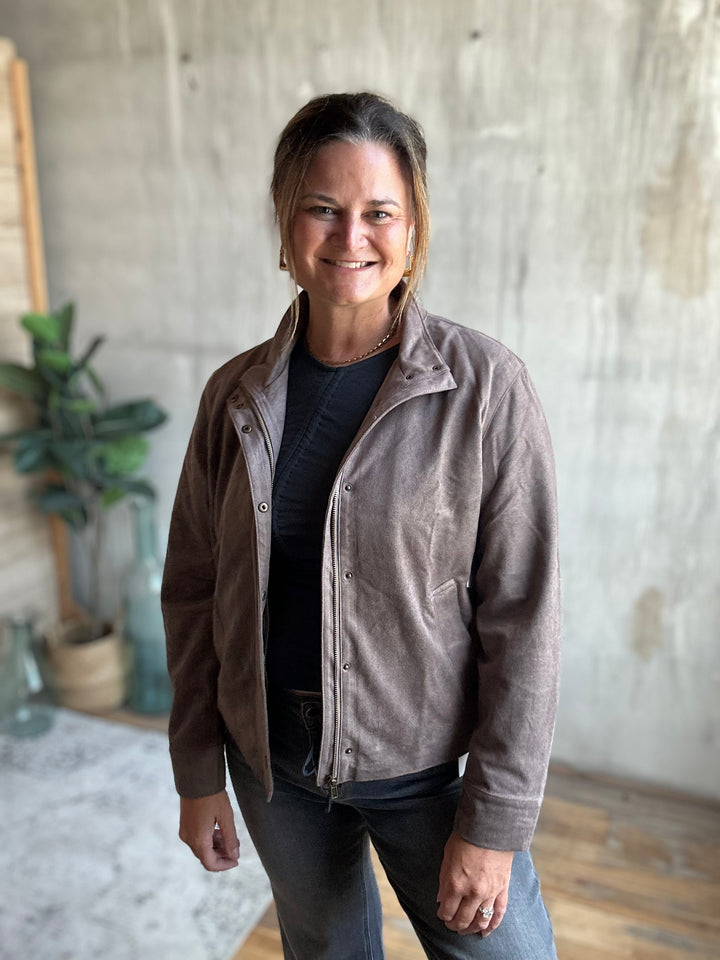 A woman wearing a brown suede button-up jacket over a black shirt and gray jeans, standing in a room with a concrete wall background.