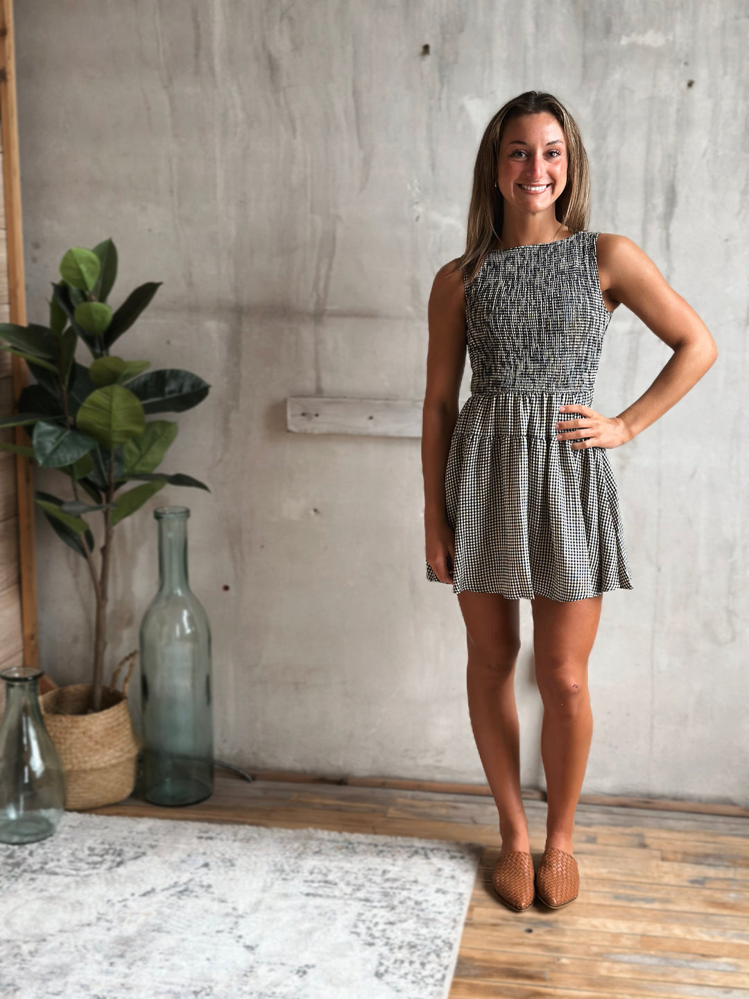 Woman in a gingham dress standing in a room with a plant and bottles.