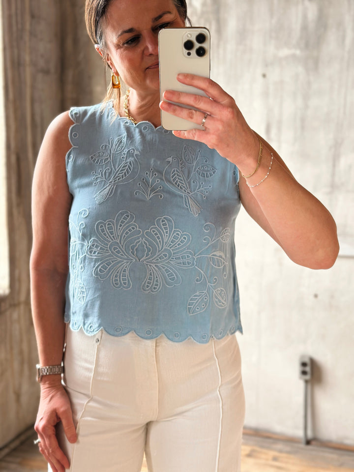 Woman taking a mirror selfie wearing a light blue top with embroidered details and white pants.