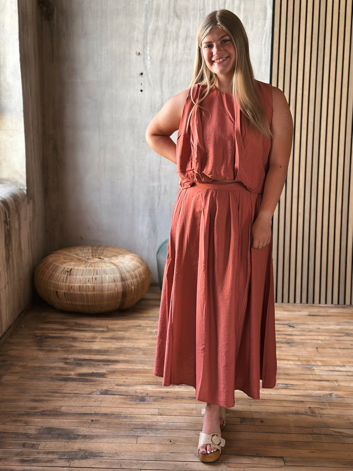 Woman in a rust-colored skirt and sleeveless top set standing in a room with wooden flooring and a textured wall.