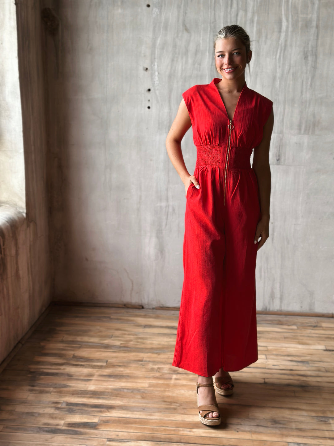 Front view of sleeveless red jumpsuit features a flattering v-neckline, a front zipper closure, and a smocked waist