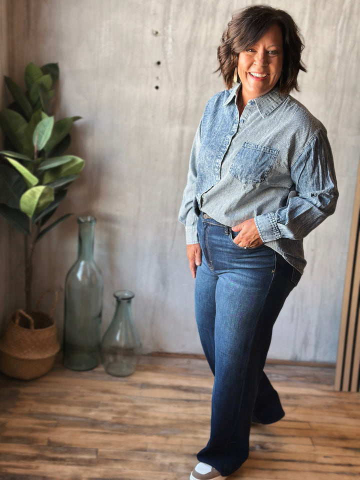 Woman wearing a denim shirt and jeans standing in a room with wooden floor and plants.