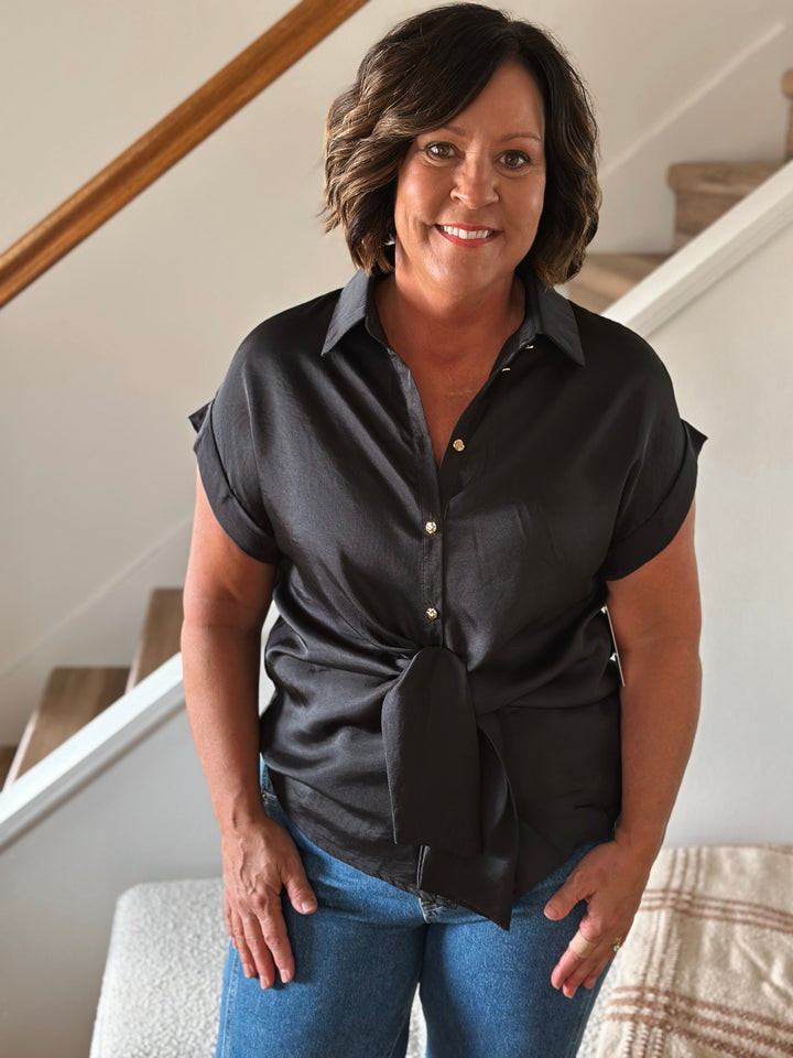Woman wearing a black short-sleeve shirt with a tie at the waist, standing indoors.