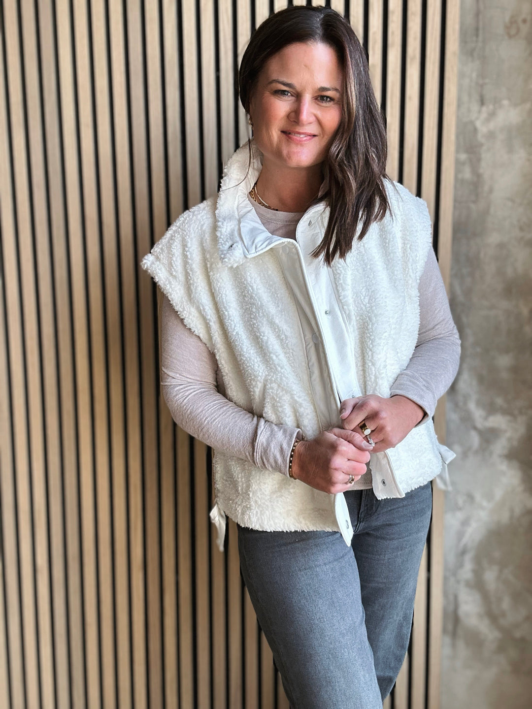 Woman wearing a white fleece vest over a light gray shirt and blue jeans, standing against a wooden paneled wall.