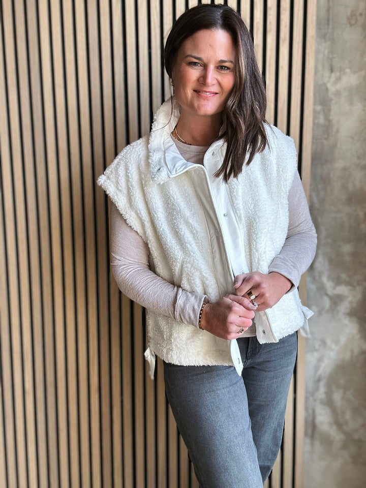 Woman wearing a white fleece vest over a light gray shirt and blue jeans, standing against a wooden paneled wall.
