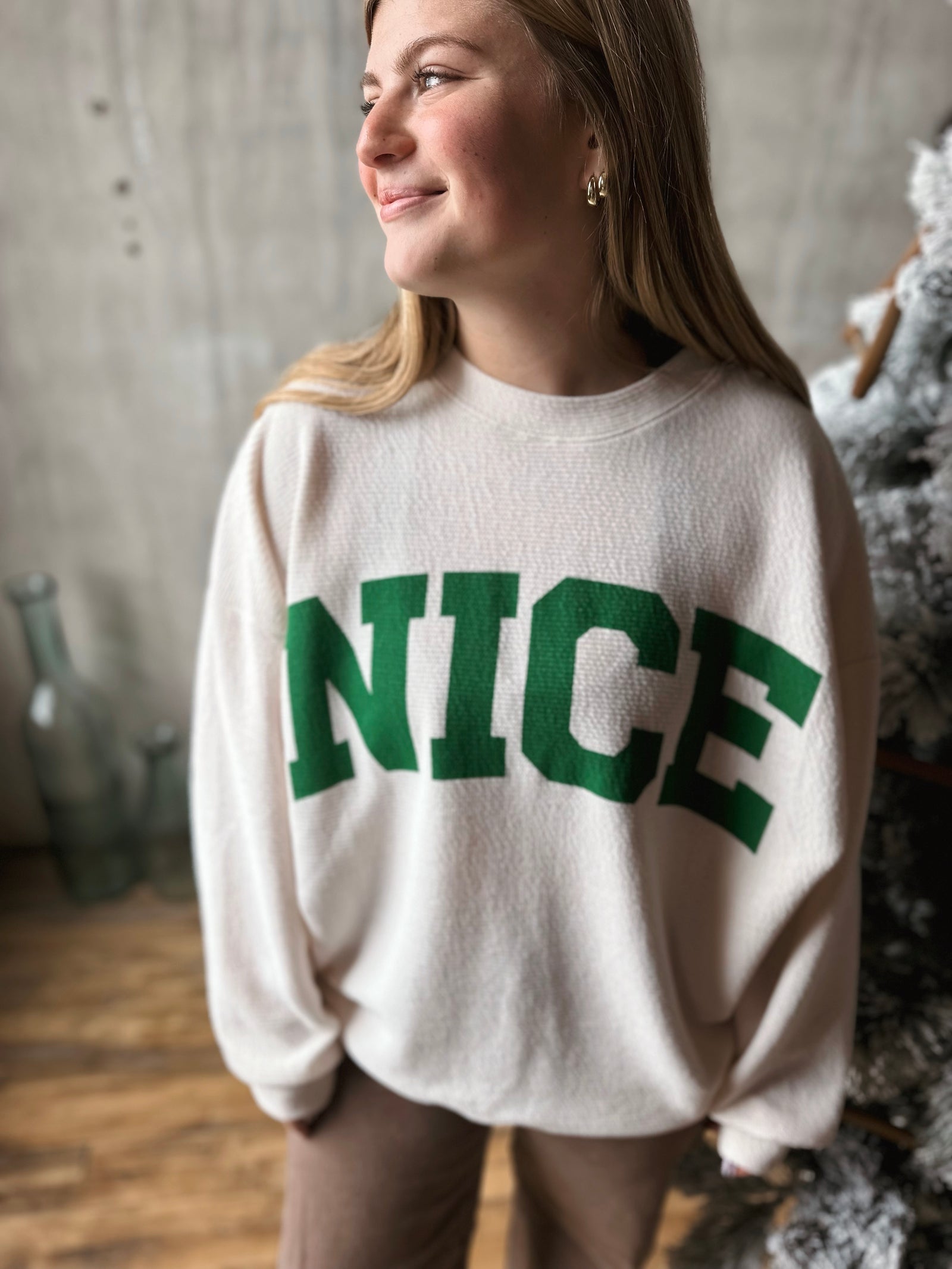 Person wearing a white sweater with 'NICE' in green letters, standing indoors.