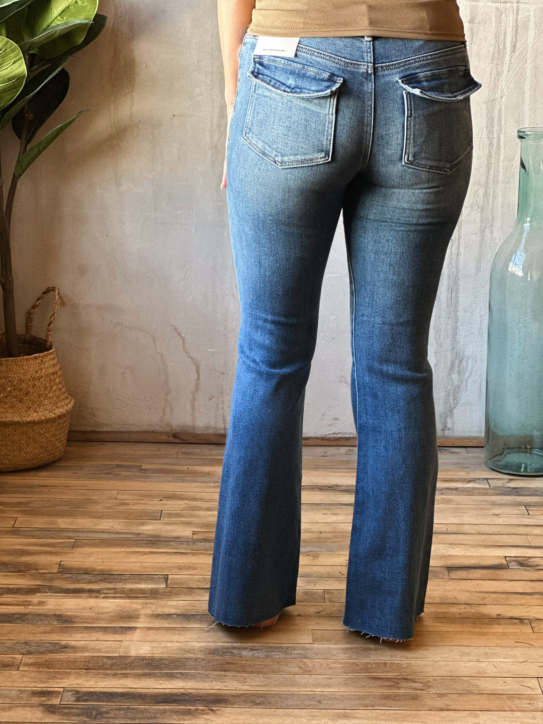 Person wearing blue flared jeans standing on a wooden floor with a plant and vase in the background.
