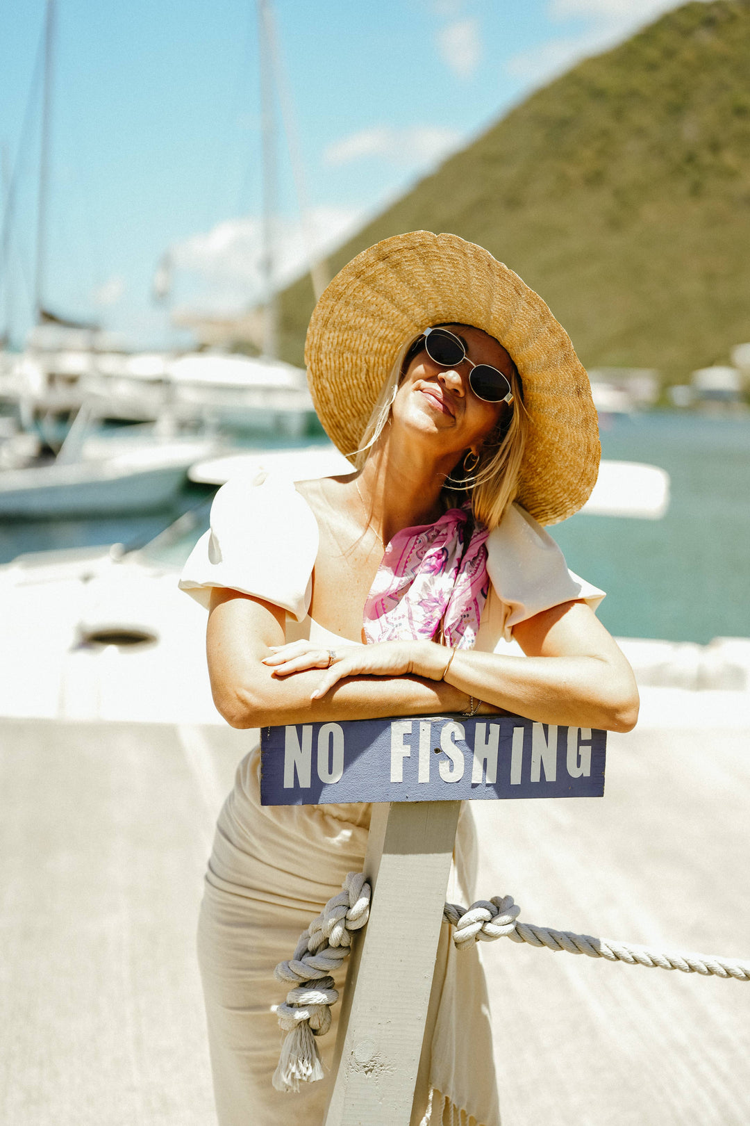 Woman in a straw hat standing next to a 'No Fishing' sign on a dock with boats and mountains in the background.