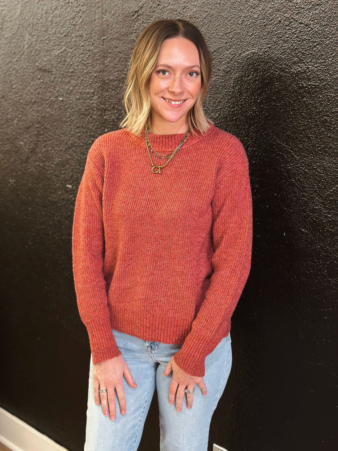 Woman wearing a rust-colored sweater and light blue jeans against a dark wall.