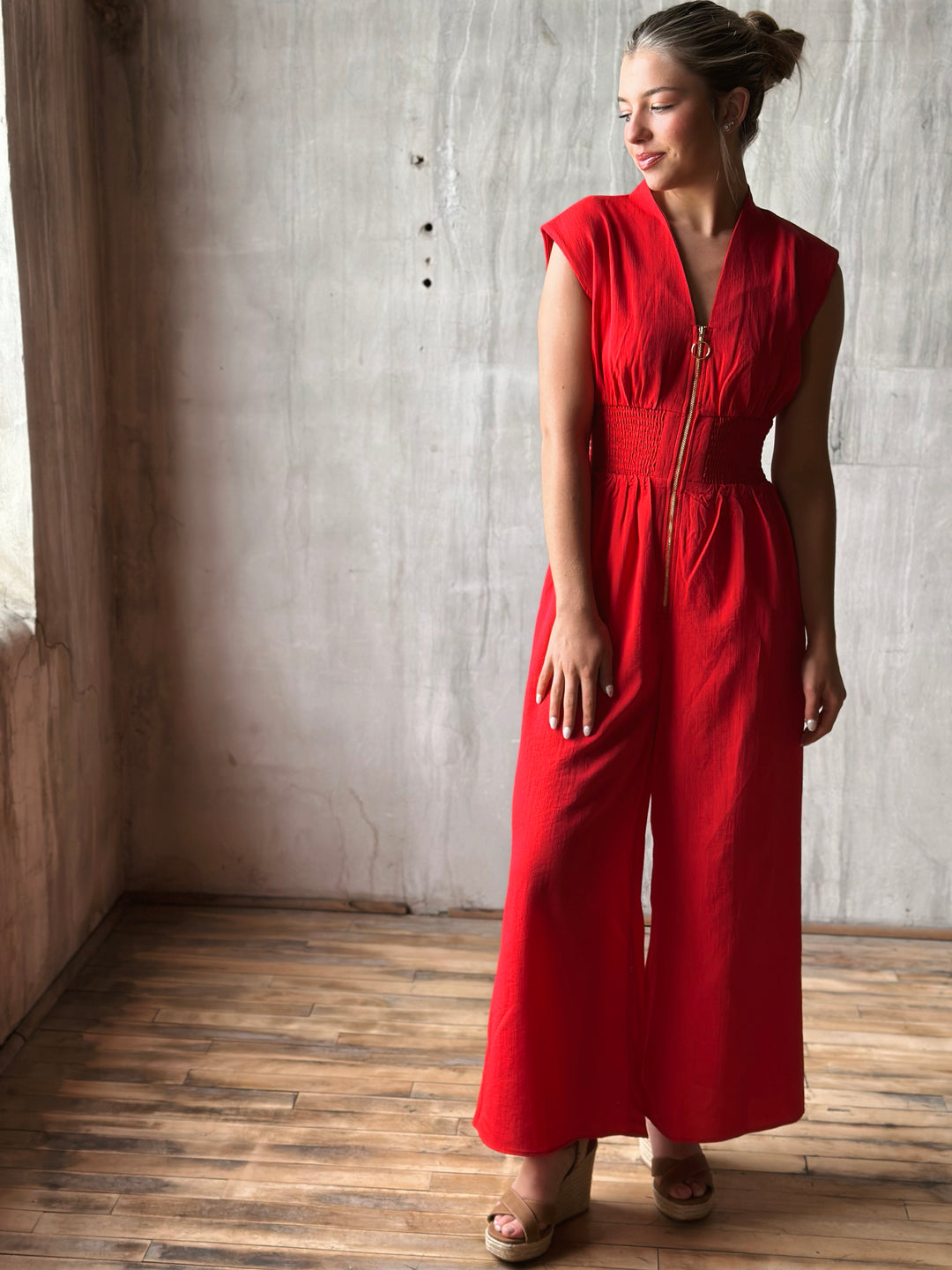 Front view of red sleeveless jumpsuit features a flattering v-neckline, a front zipper closure, and a smocked waist