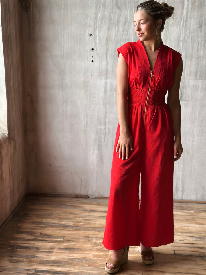 Front view of red sleeveless jumpsuit features a flattering v-neckline, a front zipper closure, and a smocked waist