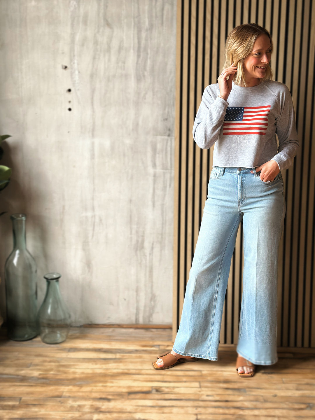 Woman wearing a gray sweater with an American flag design and light blue jeans in a room with wooden flooring and a concrete wall.