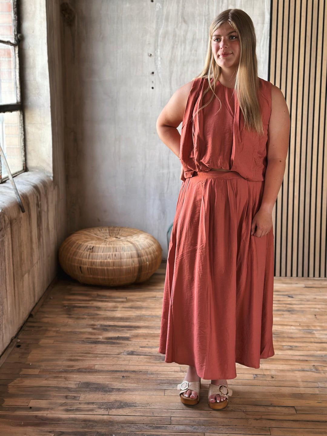Woman in a rust-colored skirt and sleeveless top set standing in a room with wooden flooring and a textured wall.