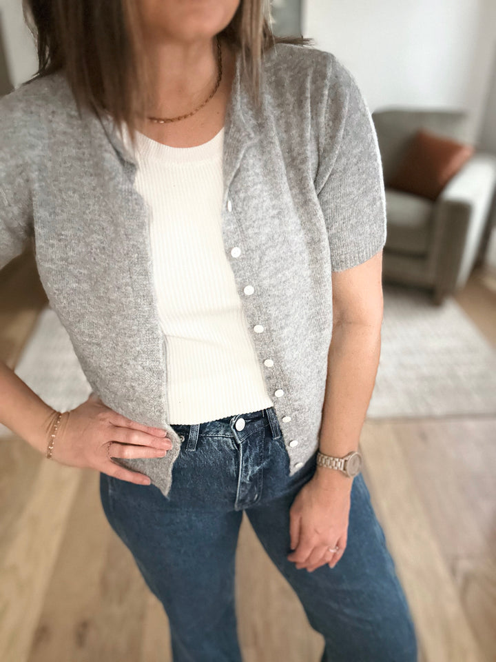 Person wearing a gray cardigan over a white shirt and blue jeans indoors.