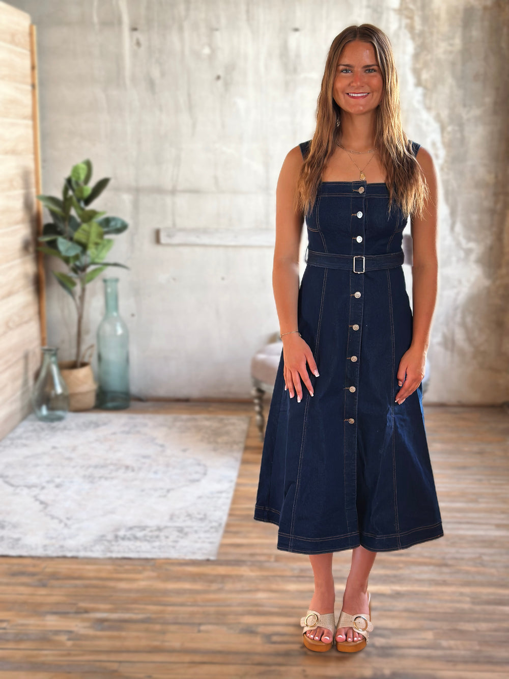 Woman wearing a dark blue denim dress with silver buttons standing in a room with a concrete wall and wooden floor.
