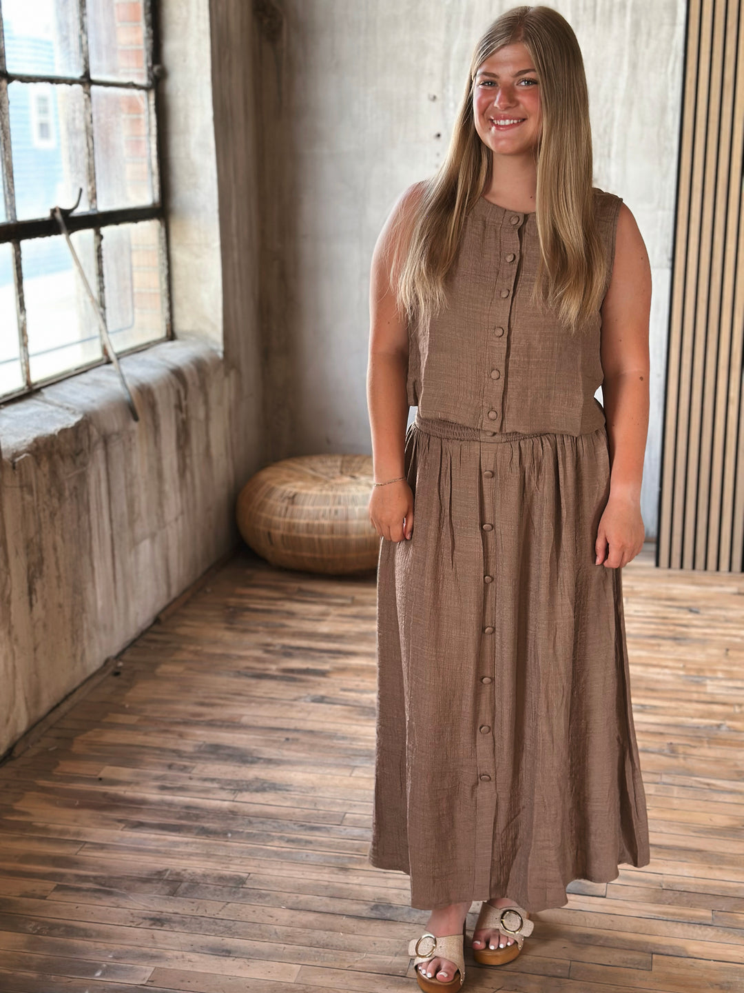 A woman wearing a brown skirt with buttons and a matching sleeveless top  standing in a room with a wooden floor and window.