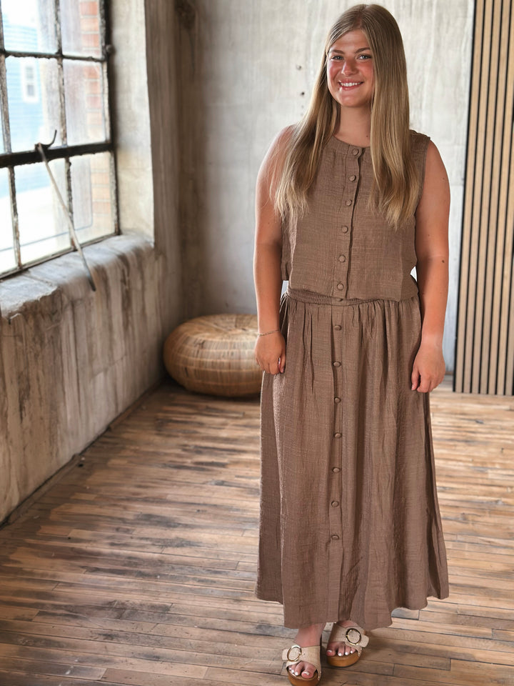 A woman wearing a brown skirt with buttons and a matching sleeveless top  standing in a room with a wooden floor and window.