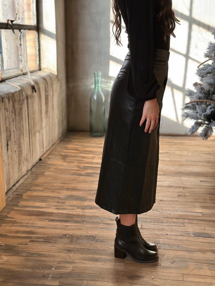 Person wearing a long black coat and boots in a room with wooden flooring and large windows.