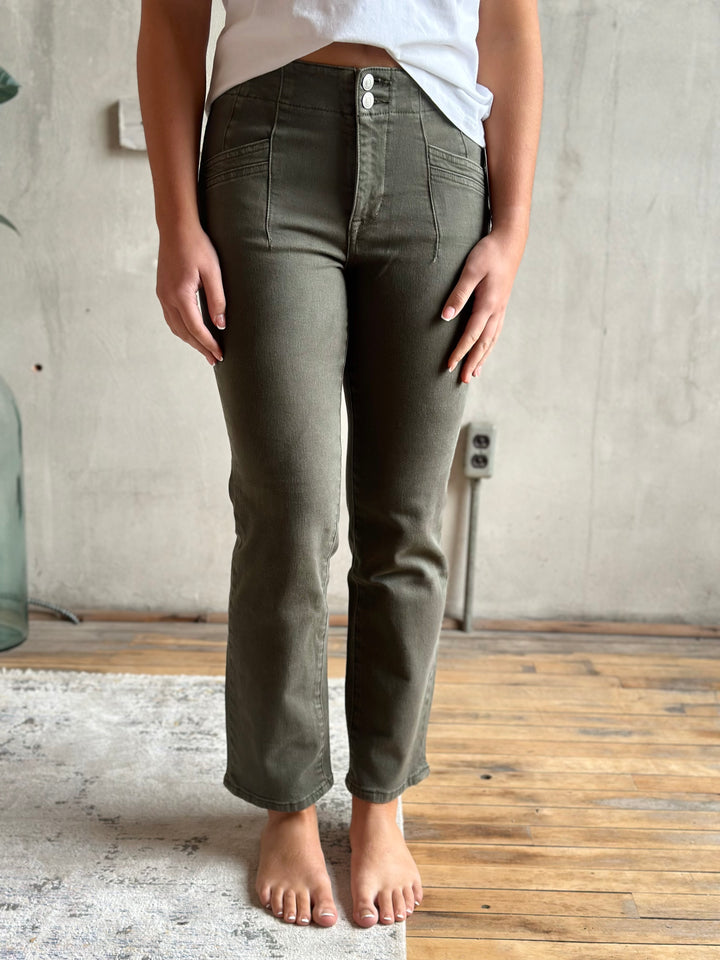 A person wearing olive green straight-leg jeans with a white top, standing barefoot on a light rug in a room with a concrete wall and wooden floor.