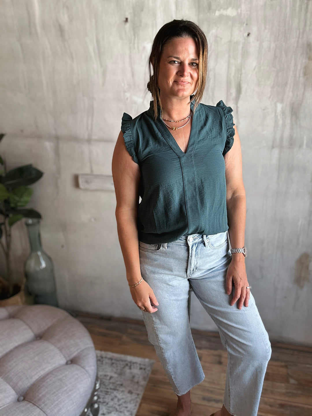 Woman wearing a dark green sleeveless top with ruffles on the sleeves and light blue jeans.