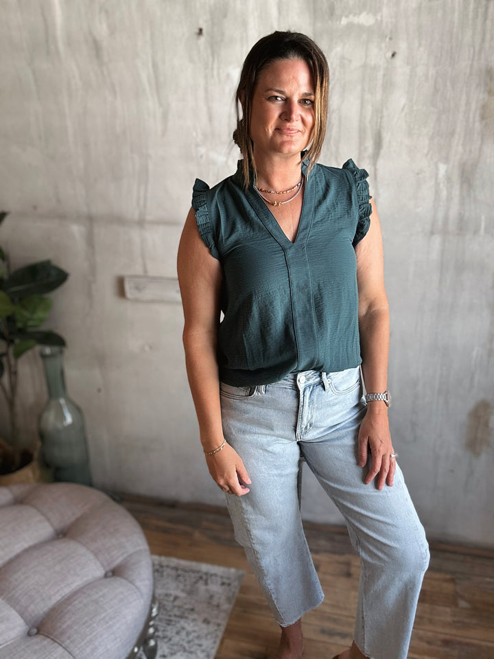 Woman wearing a dark green sleeveless top with ruffles on the sleeves and light blue jeans.