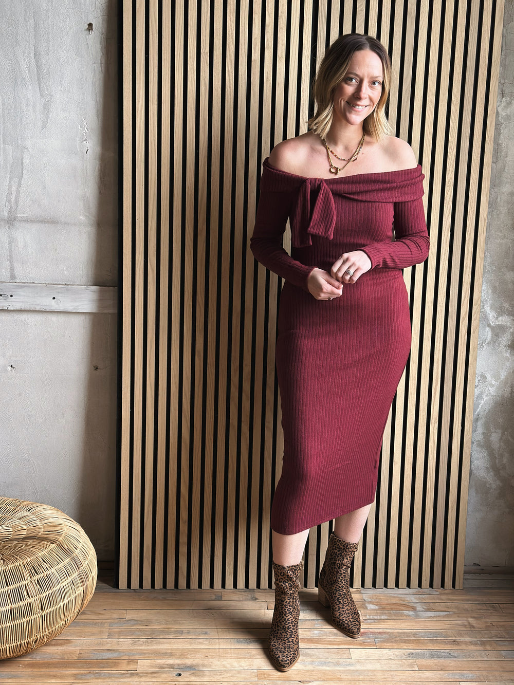 Woman wearing a burgundy off-shoulder dress standing against a wooden panel wall.
