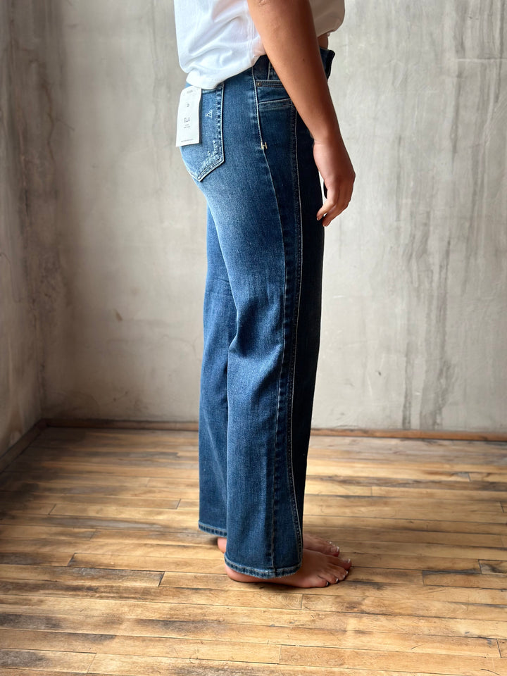A person wearing a pair of wide-leg blue denim jeans with a white top, standing barefoot on a wooden floor against a textured concrete wall.