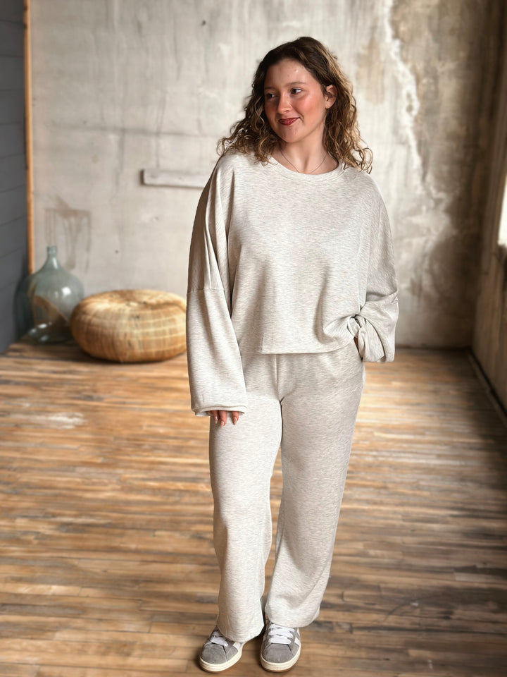 Woman wearing a light heather grey sweat set standing in a room with wooden flooring and a textured wall.