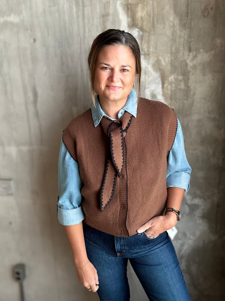 A woman wearing a brown ribbed knit vest over a light blue button-up shirt, paired with blue jeans, standing against a textured concrete wall.