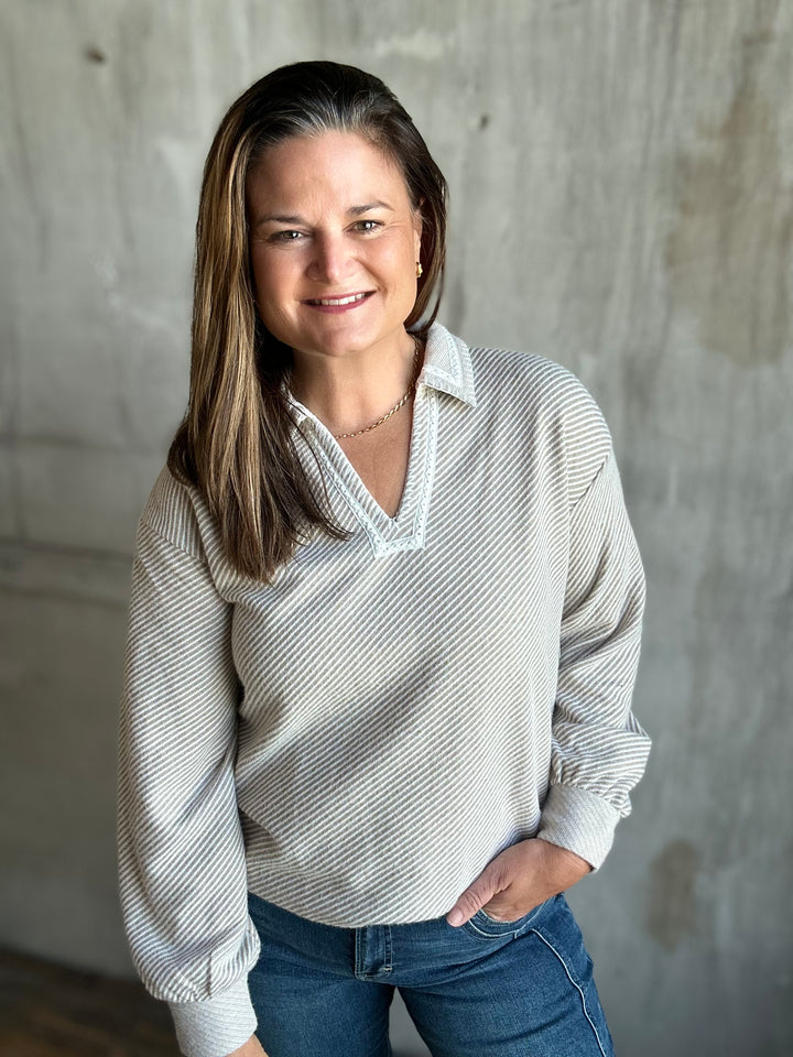 A woman with long brown hair smiling, wearing a beige and white striped long-sleeve knit polo shirt with blue jeans, standing against a textured concrete wall.