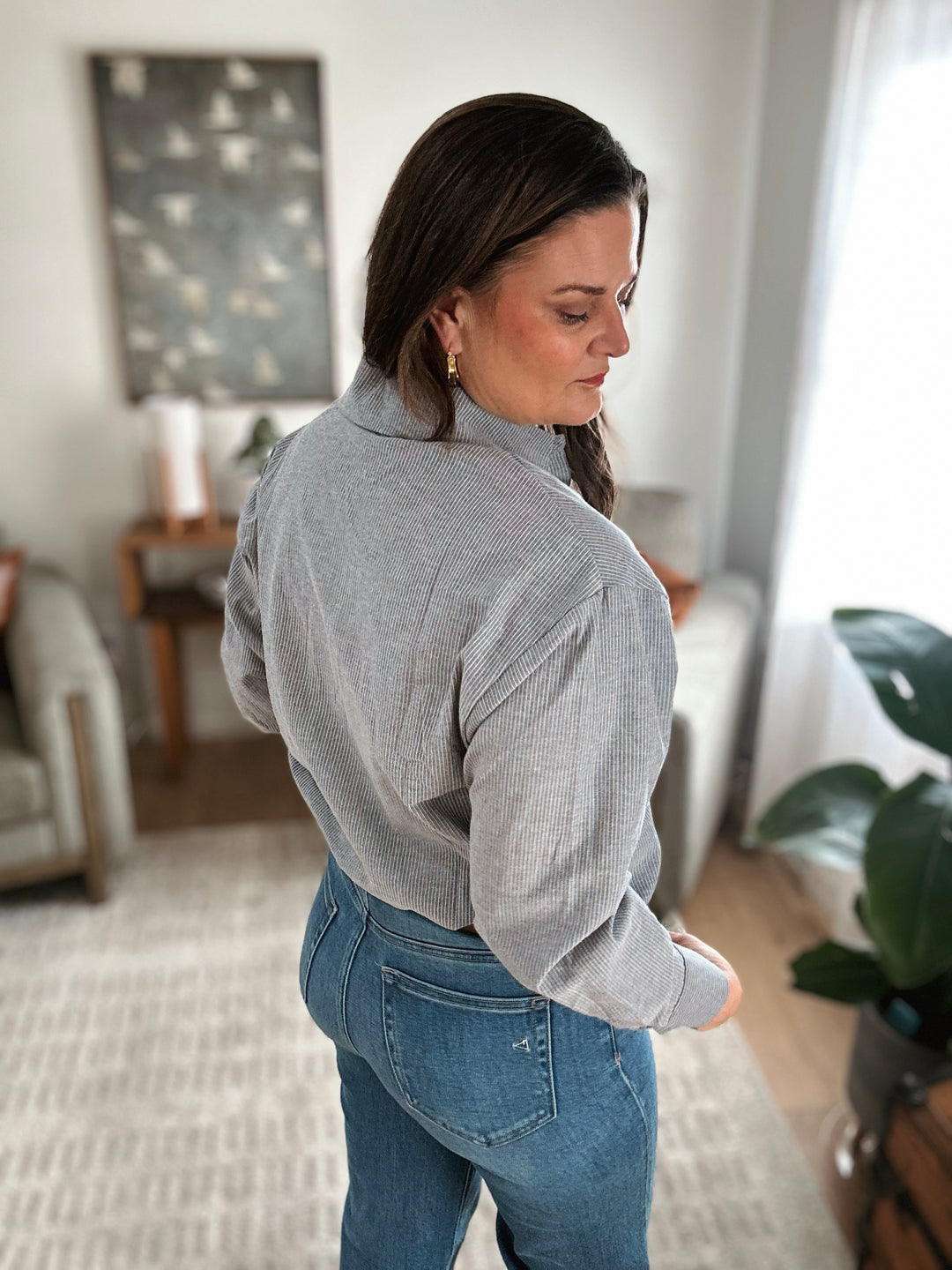 A woman wearing a gray sweater and blue jeans in a room with a plant and furniture.