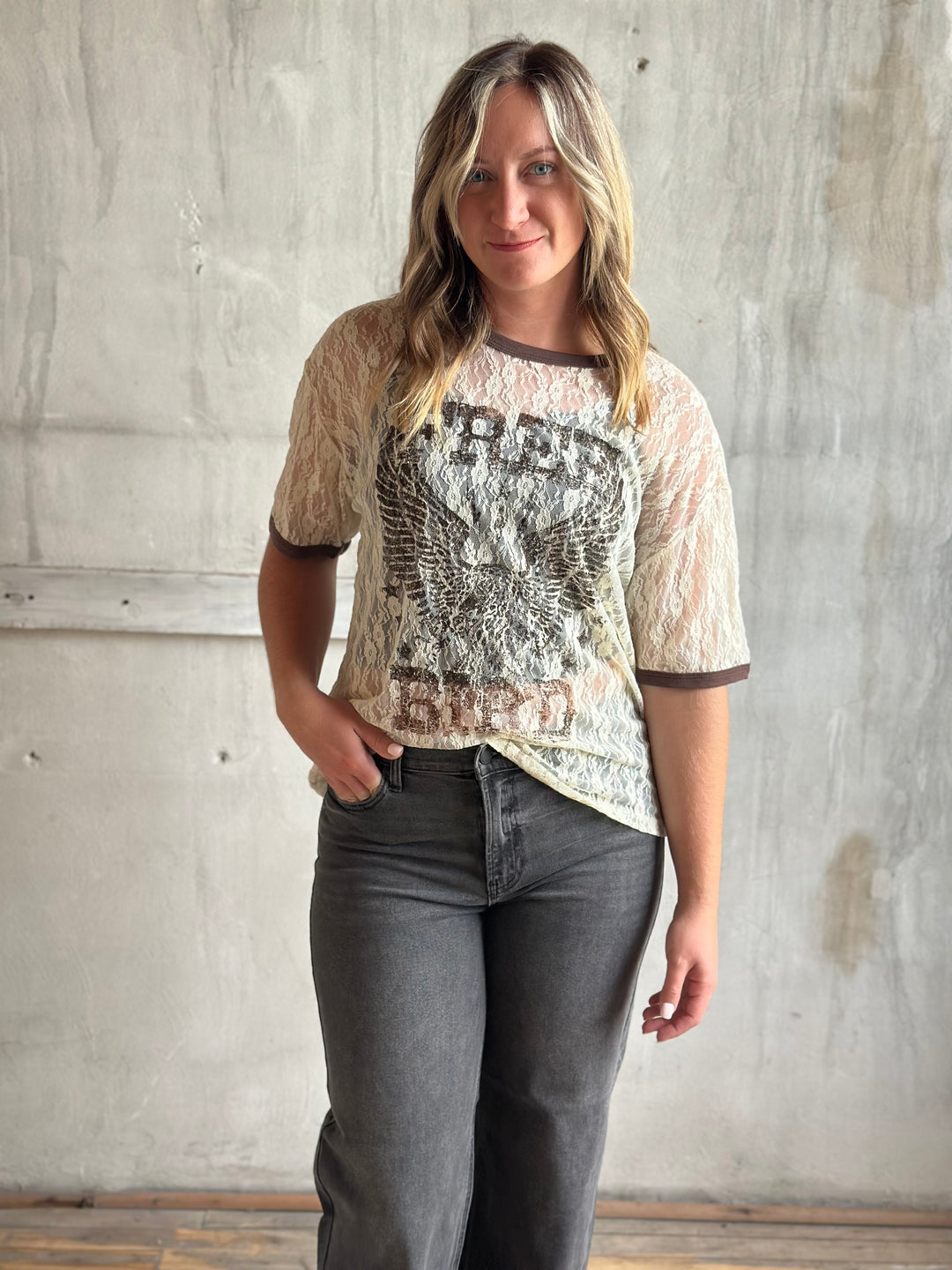 Woman wearing a cream lace graphic tee with an eagle print and brown trim, with gray jeans