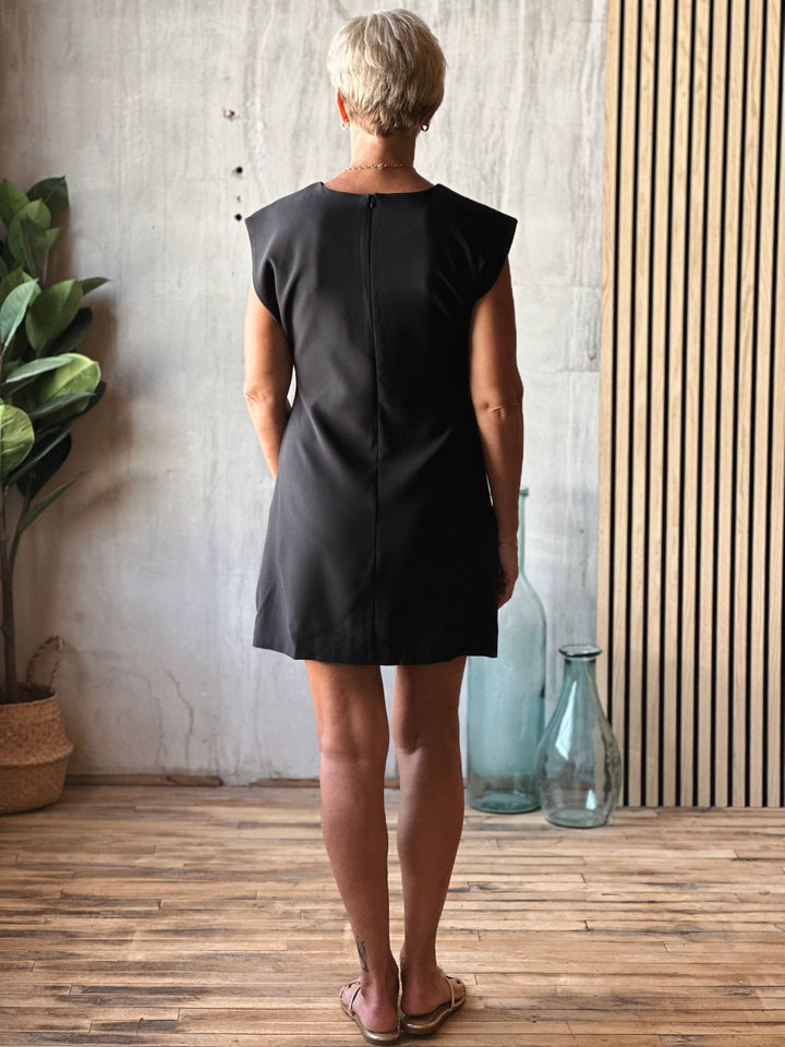 Person wearing a black dress standing in a room with a plant and decorative bottles.