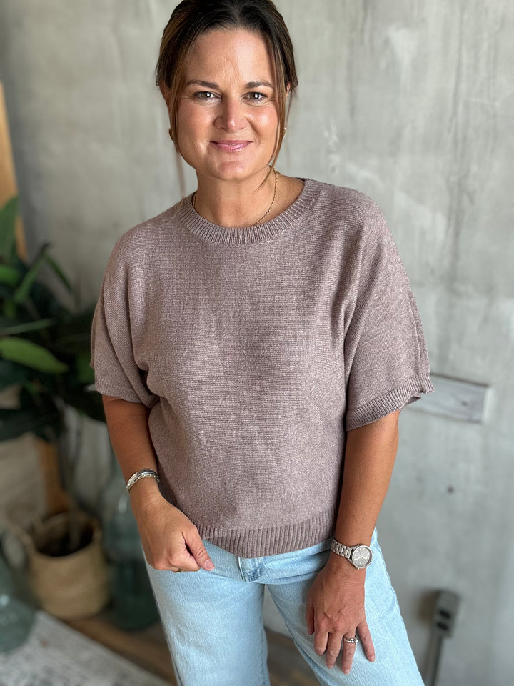 A woman wearing a short-sleeve knit  sweater in a dusty mauve-brown color, paired with light blue jeans.